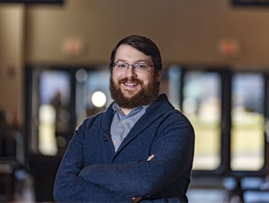 Travis Loof stands with his arms crossed, a wide smile travels across his face. He is standing in the hallway of a brightly lit Al Neuharth Building.