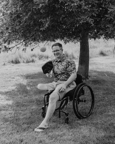 A person in a wheelchair sits under a tree and holds a pickleball paddle. 