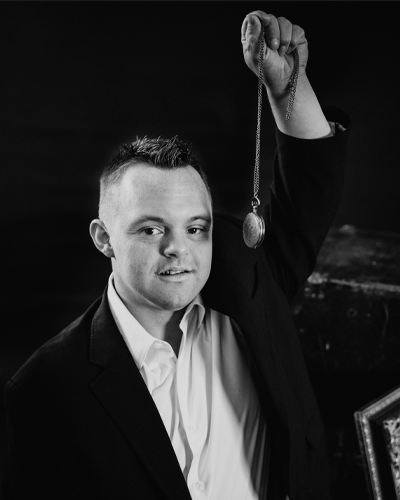 A person in a suit coat and dress shirt holds up a locket necklace and smiles.