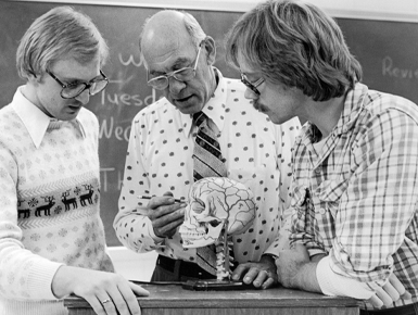 A black and white photo of a professor showing two students a model of a human skull.