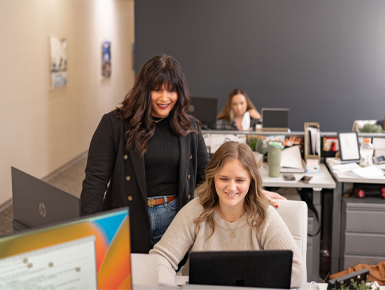 Addie stands over one of her employees shoulders and looks at a computer screen while smiling.