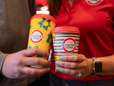 Two hands hold Scooters Coffee cups and touch the cups together..